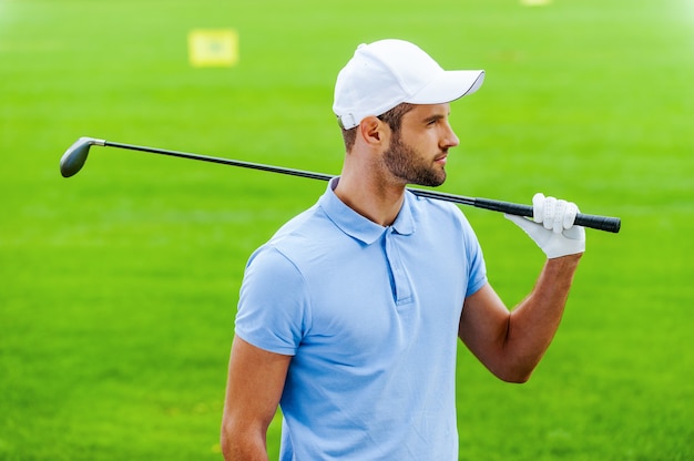 Thinking about next shot. Confident male golfer carrying driver on shoulder and looking away while standing on golf course