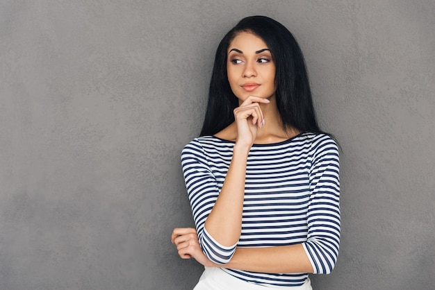 Thinking about solution. Beautiful young African woman looking away and holding hand on chin while standing against grey background