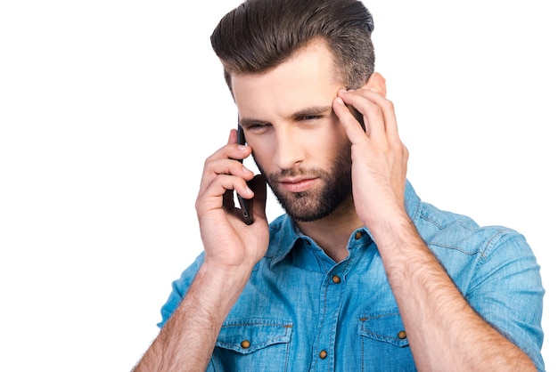 Thinking about solution. Concentrated young handsome man in jeans shirt talking on mobile phone while standing against white background