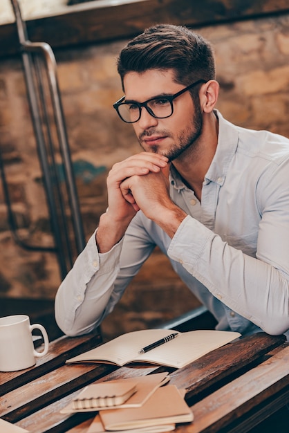 Thinking about solution. Handsome thoughtful young man keeping hands clasped and looking away while sitting at his working place