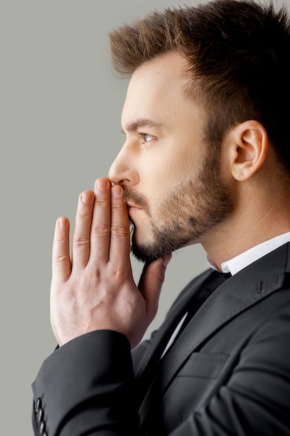 Thinking about solutions. Side view of young man in formalwear holding hands clasped and looking away while standing against grey background