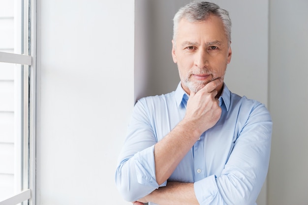 Photo thinking about solutions. thoughtful grey hair senior man in shirt looking away while standing near the window