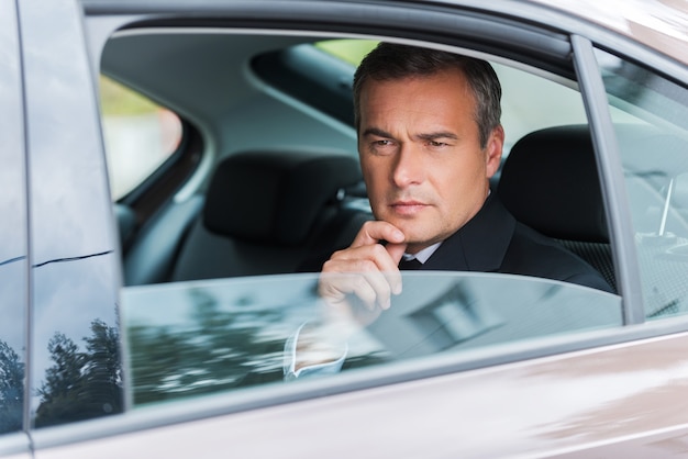 Thinking about solutions. Thoughtful mature businessman holding hand on chin and looking away while sitting on the back seat of a car