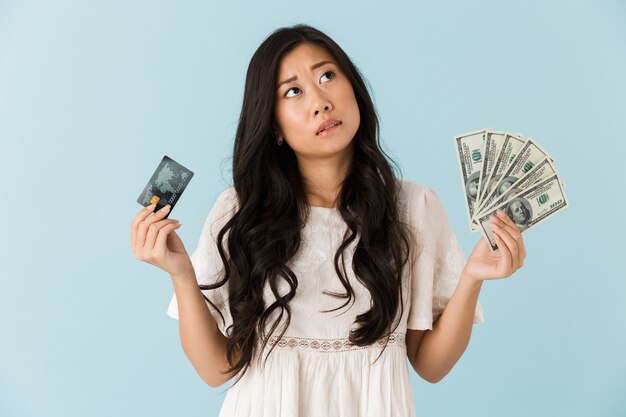 Thinking asian beautiful woman isolated over blue wall holding credit card and money