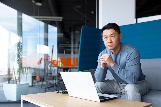 생각하는 아시아 남자는 현대 사무실의 컴퓨터에서 일하고 프리랜서는 중요한 일을 해결하는 데 집중합니다