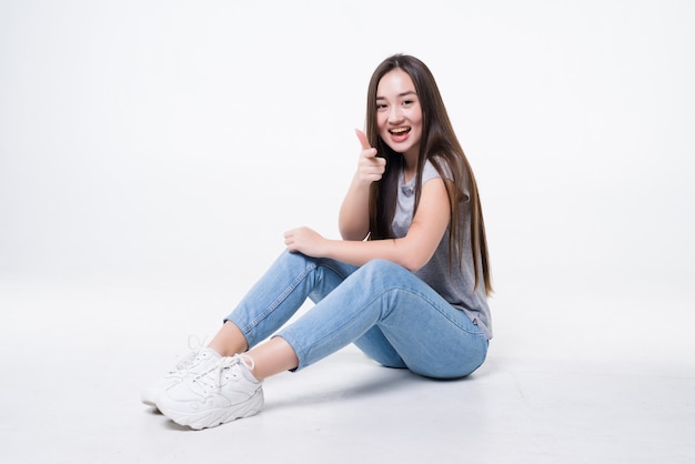 Thinking asian woman sitting on floor pointed on you isolated on white wall.