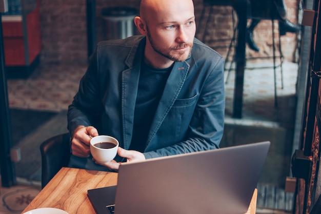 Thinking attractive adult successful bald bearded man in suit with laptop in cafe