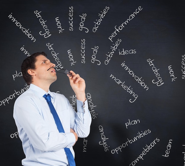 Thinking businessman biting glasses against blackboard with business buzzwords