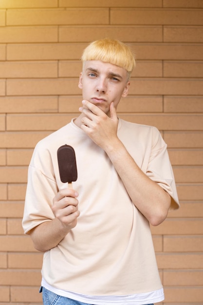 A thinking Caucasian man dressed in a beige Tshirt and holding a chocolate ice cream on a stick