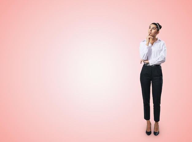 Концепция мышления с задумчивой деловой женщиной в белой рубашке и темных брюках, стоящей в полный рост на абстрактном светло-розовом фоне стены с пустым местом для текста, макет