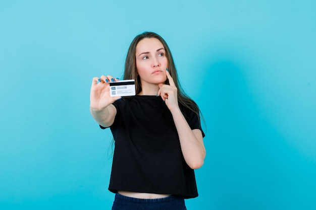 Thinking girl is holding forefinger on cheek and showing credit card to camera on blue background