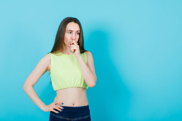 Thinking girl is putting one hand on mouth and other on waist on blue background