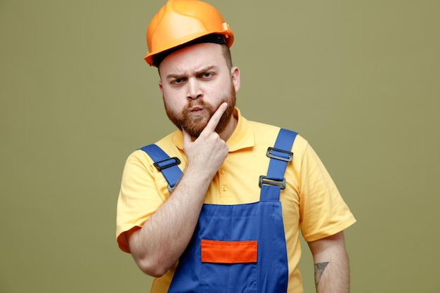 Thinking grabbed chin young builder man in uniform isolated on green background