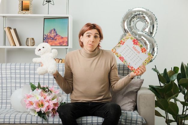 Thinking handsome guy on happy women day holding teddy bear with calendar sitting on sofa in living room