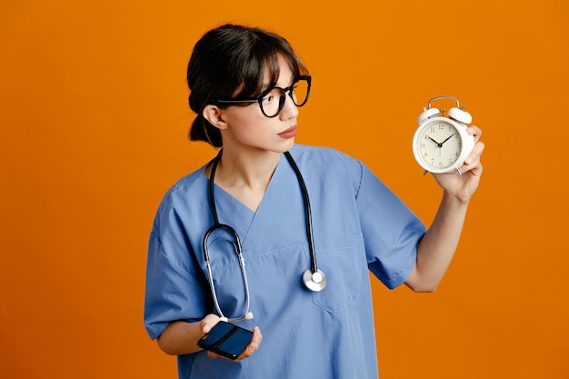 Thinking holding alarm clock speaks on the phone young female doctor wearing uniform fith stethoscope isolated on orange background