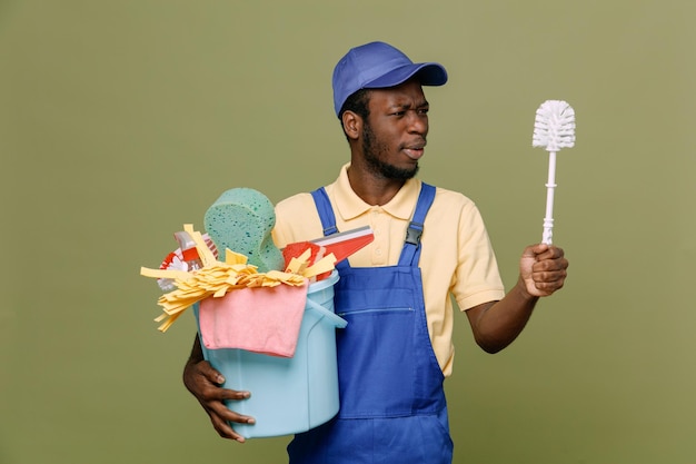 Thinking holding bucket of cleaning tools and looking at cleaning brush in his hand young africanamerican cleaner male in uniform with gloves isolated on green background