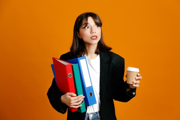 Thinking holding folders with coffee cup young beautiful female wearing black jacket isolated on orange background