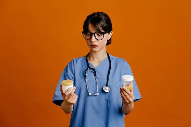 Thinking holding pill containers young female doctor wearing uniform fith stethoscope isolated on orange background