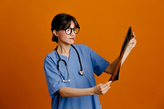 Thinking holding xray young female doctor wearing uniform fith stethoscope isolated on orange background