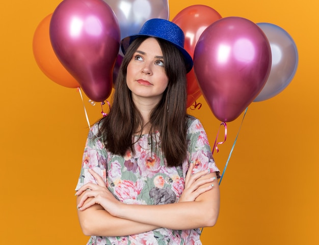 Thinking looking side young beautiful girl wearing party hat standing in front balloons crossing hands 