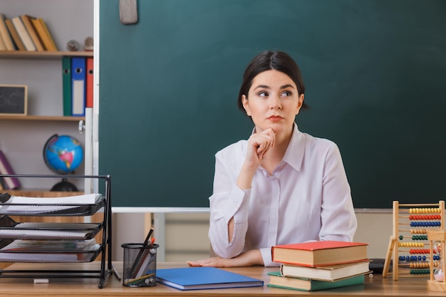 thinking looking at side young female teacher sitting at desk with school tools in classroom