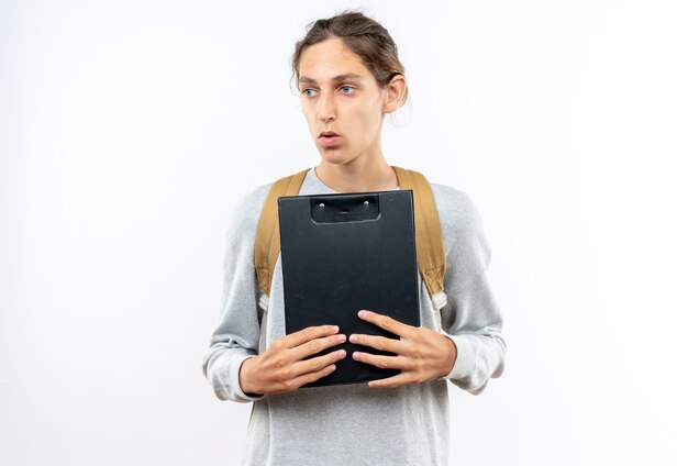 Thinking looking side young guy student wearing backpack holding clipboard isolated on white wall