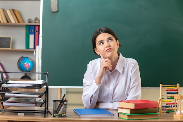 Thinking looking up putting hand on cheek young female teacher sitting at table with school tools in classroom