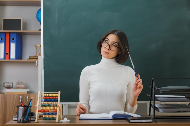 thinking looking at up young female teacher wearing glasses putting pointer on head sitting at desk with school tools on in classroom