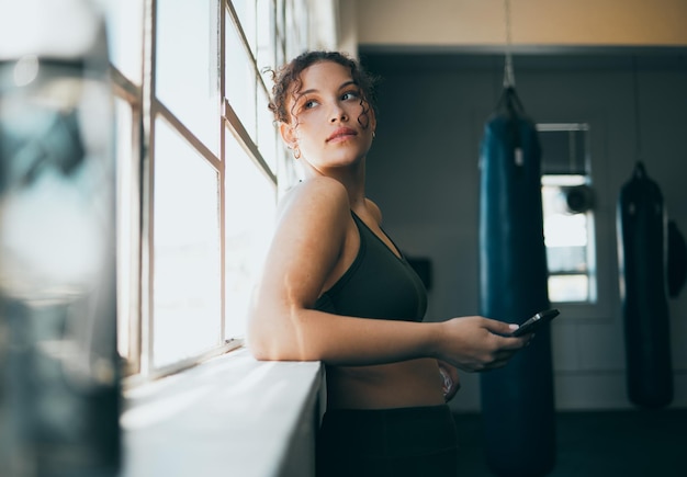 Photo thinking phone and fitness with a sports woman by a window standing in the gym during an exercise workout health idea and a female athlete using social media or an app to track her training