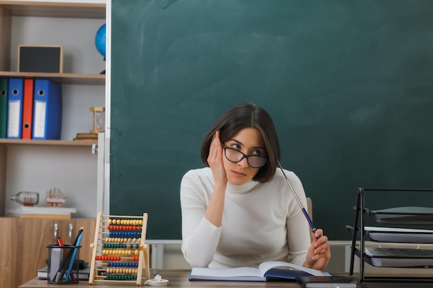 thinking putting hand on cheek young female teacher wearing glasses holding pointer sitting at desk with school tools on in classroom