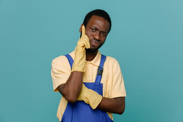 Pensando di mettere la mano sul viso giovane maschio pulitore afroamericano in uniforme con guanti isolati su sfondo blu