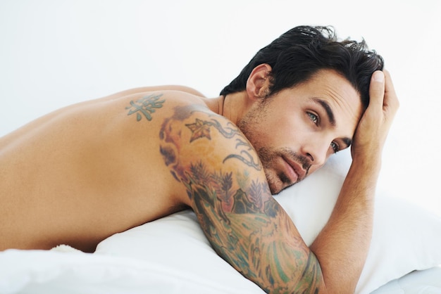 Thinking relax and sexy with a topless man on a bed lying in studio on a white background Tattoo idea and shirtless with a handsome young male model posing in a bedroom for sensuality or desire