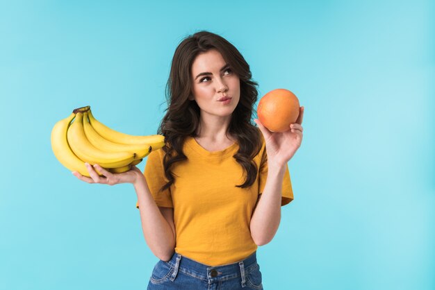 바나나와 오렌지 사이를 선택 하는 파란색 벽 위에 고립 된 포즈를 생각 심각한 젊은 예쁜 여자.