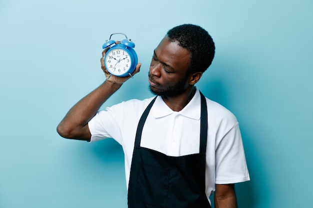 Thinking young african american barber in uniform holding and looking at alarm clock isolated on blue background