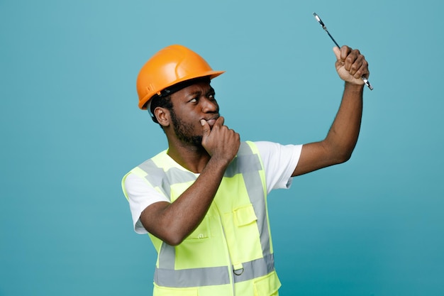 Thinking young african american builder in uniform holding and looking at open end wrench isolated on blue background