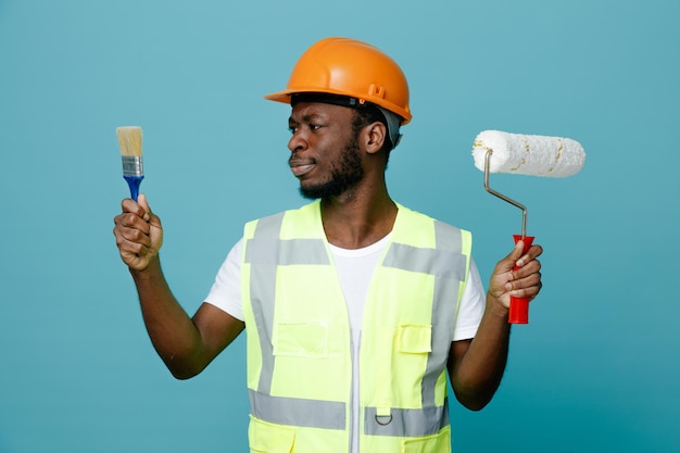 Thinking young african american builder in uniform holding and looking at roller brush with paint brush isolated on blue background