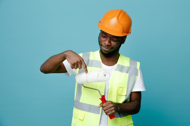 thinking young african american builder in uniform holding roller brush isolated on blue background