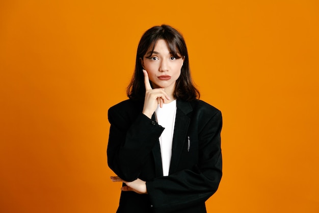 Thinking young beautiful female wearing black jacket isolated on orange background