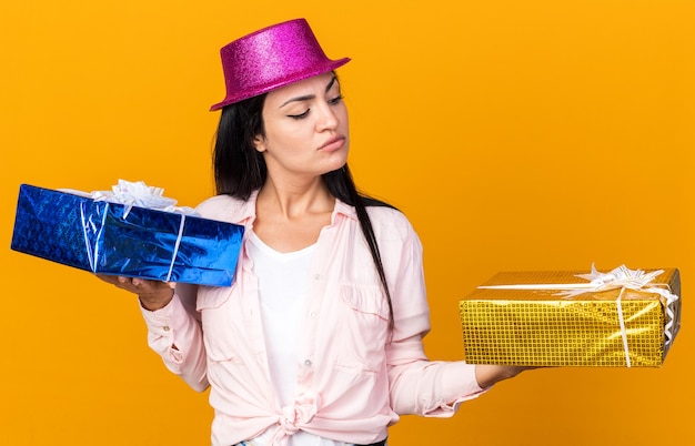 Foto pensando a una giovane bella ragazza che indossa un cappello da festa che tiene e guarda scatole regalo isolate sulla parete arancione