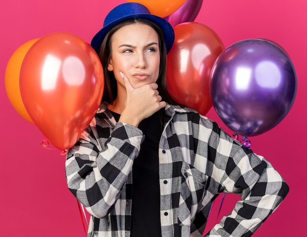 Thinking young beautiful girl wearing party hat standing in front balloons grabbed chin 