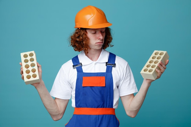 Thinking young builder man in uniform holding and looking at bricks isolated on blue background