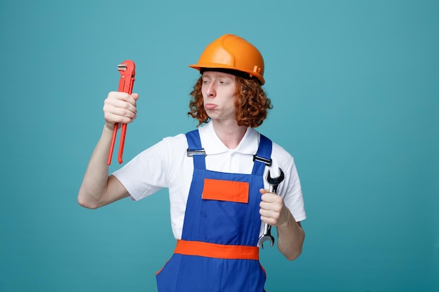 Thinking young builder man in uniform holding and looking at wrenches isolated on blue background