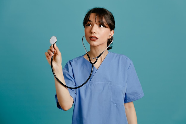 青い背景に分離された均一なフィス聴診器を身に着けている若い女性医師を考える