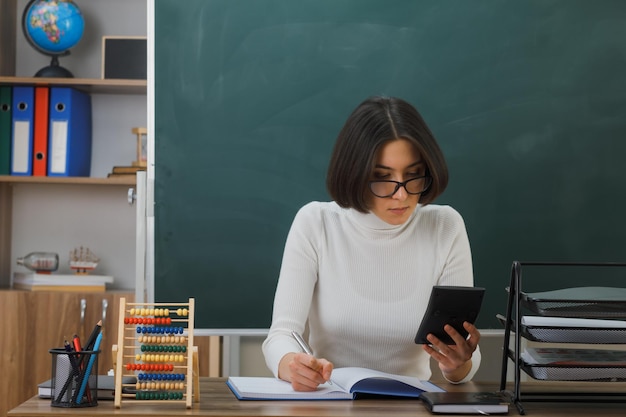 thinking young female teacher wearing glasses holding calculator writting sitting at desk with school tools on in classroom