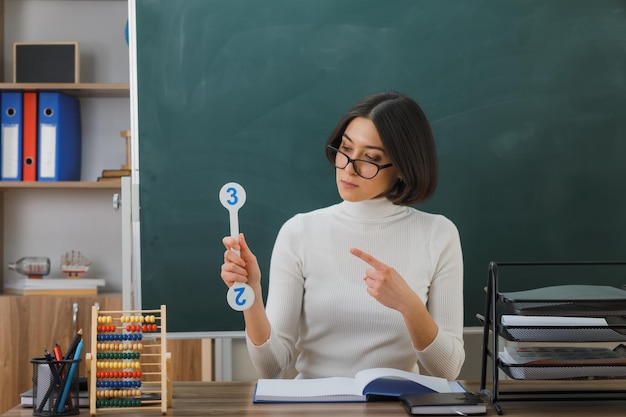 thinking young female teacher wearing glasses holding and points at number fan sitting at desk with school tools on in classroom