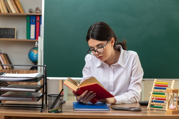 Thinking young female teacher wearing glasses reading book sitting at table with school tools in classroom
