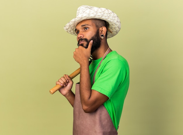 Thinking young gardener afro-american guy wearing gardening hat holding rake grabbed chin 