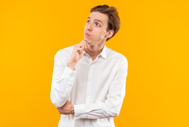 Pensando giovane bel ragazzo che indossa una camicia bianca afferrò il mento isolato sul muro arancione