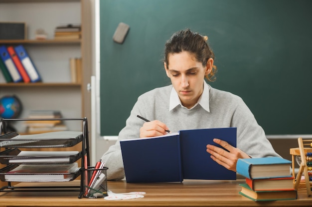 thinking young male teacher holding notebbok with pencil sitting at desk with school tools on in classroom