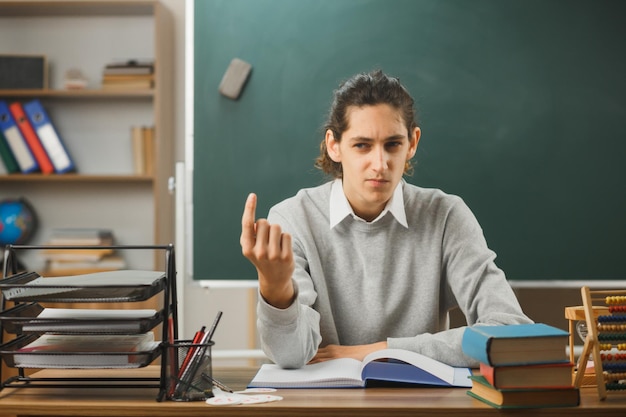 思考の若い男性教師は教室で学校のツールをオンにして机に座って上を指しています